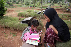 Adinda Rahma Barokah, 7, squats near a tombstone while doing her school assignment in Padurenan cemetery in Bekasi, West Java, on Oct. 15. She read the questions on her mother’s phone and wrote down the answers in her notebook. As her school -- SD Wanarasi 15 state elementary school -- is closed due to the outbreak, Adinda has been studying in the cemetery where her mother sells snacks and drinks. JP/P.J.Leo